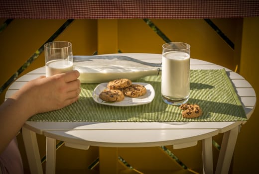 Glass of milk held in a woman's hand and a plate with cookies, on a white round table, in a warm sunny morning.