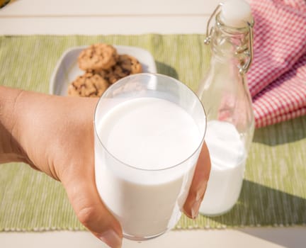 Food photography with a glass of milk held in a woman's hand, the bottle of milk and cookies lying on the table in the background.