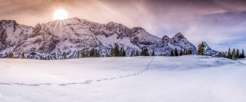 Winter landscape with Austrian alps peaks, a single trail of footsteps on snow, under a dramatic and colorful sky.