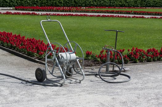 Watering Equipment with  Pump and Hose Reel on Gravel Track near Flower Bed with Red Flowers and Green Grass Outdoors