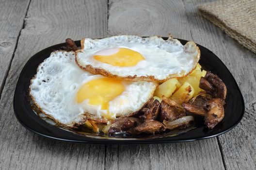 Fried eggs with potatoes and mushrooms on a black plate and a rough grey Board