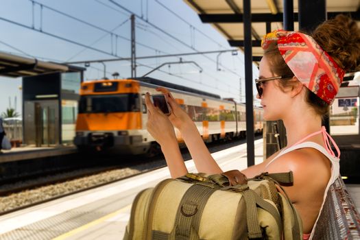 Young sexy woman backpacker sitting on bench on railway station and charting on smart phone. Travel concept.