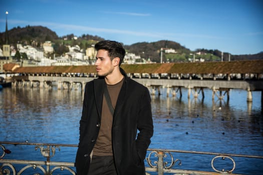 Handsome man standing near metal fence in Lausanne. Famous wooden bridge and Water Tower on background.