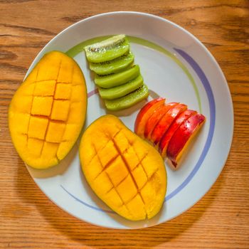 a plate of sliced multicolored ​​fruit, with mango kiwi and peach on wooden table
