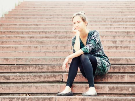 Sad lonely young woman sitting on steps. Portrait of serious girl with facial expression outdoors.