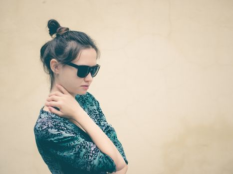 Fashionable serious young woman in black sunglasses posing near wall with copy space