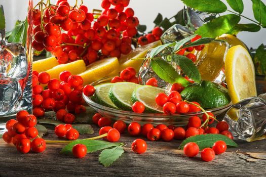 Mountain ash berries on branches with leaves and the cut citrus fruits with ice pieces on a wooden table