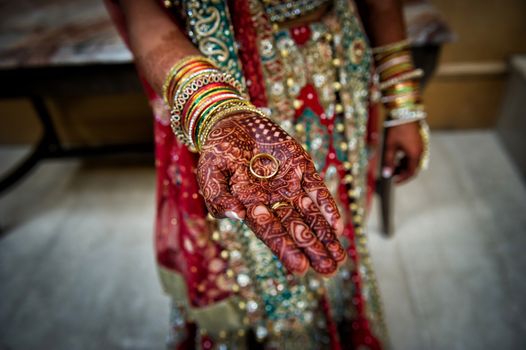 Image of an Indian bride extending hand with henna tattoos, holding groom's wedding ring toward camera