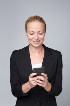 Beautiful young caucasian businesswoman in business attire using smart phone application. Studio portrait shot on grey background.