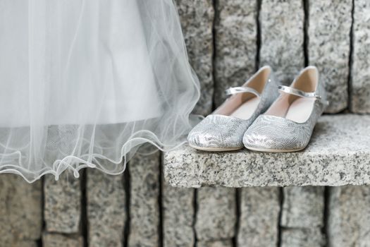 A child's wedding dress hanging up 
with silver shoes beside on granite background.