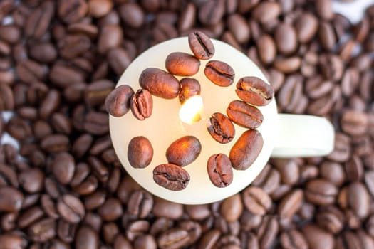 Still life of cup candle with warm flame in scattered roasted coffee beans