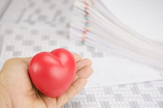 Red heart put in hand of man on finance account have blur pile overload paperwork of receipt and report with colorful paperclip as background.
