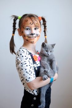 Sweet portrait of a cute little girl with face painted like a leopard holding a gray kitten