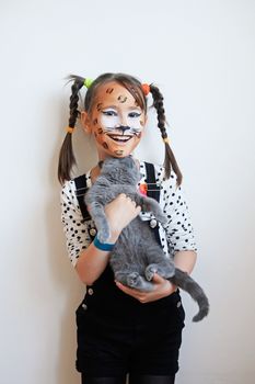Sweet portrait of a happy little girl with face painted like a leopard holding a gray kitten