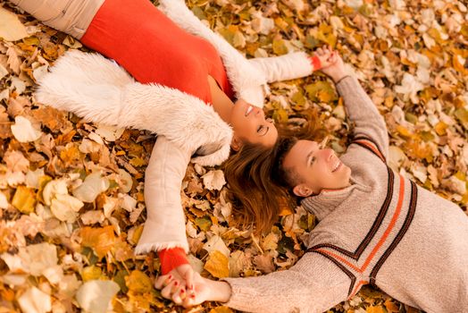 Beautiful smiling couple enjoying in sunny forest in autumn colors. They are lying on the falls leaves.