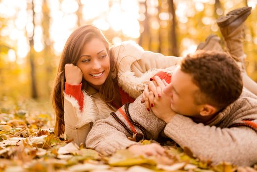 Beautiful smiling couple enjoying in sunny forest in autumn colors. They are lying on the falls leaves and having fun.