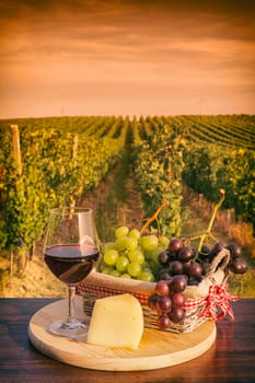 Glass of red wine with grapes in a basket and cheese in front of a vineyard at sunset