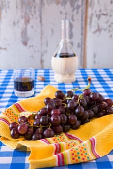 Bunch of red grapes and a glass of red wine with a wine flask on background over a checkered tablecloth
