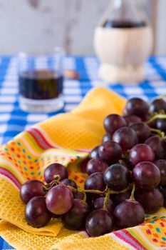 Closeup of a bunch of red grapes and a glass of red wine with a wine flask on background over a checkered tablecloth