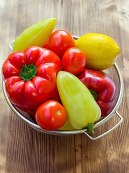 Vegetable still life of red and green peppers, tomatoes and lemon in sieve bowl on wooden board background