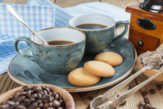 Two blue cups of black coffee, cookies and sugar pieces surrounded by checkered linen cloth and coffee beans on old wooden table