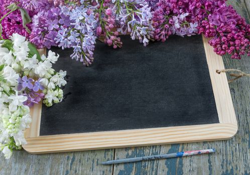 Flower border composed of lush multicolored bunches of lilac flowers around of empty black chalkboard on the old wooden background