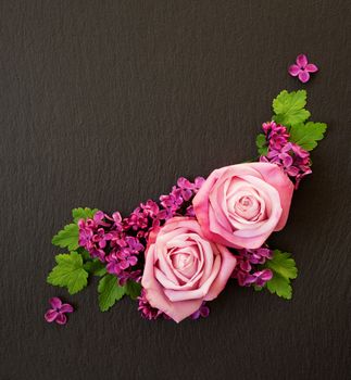 Decorative composition consisting of pink roses, violet lilac flowers and green leaves on a black slate. Flat lay, top view, overhead view