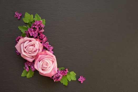 Decorative composition consisting of pink roses, violet lilac flowers and green leaves on a black slate. Flat lay, top view, overhead view