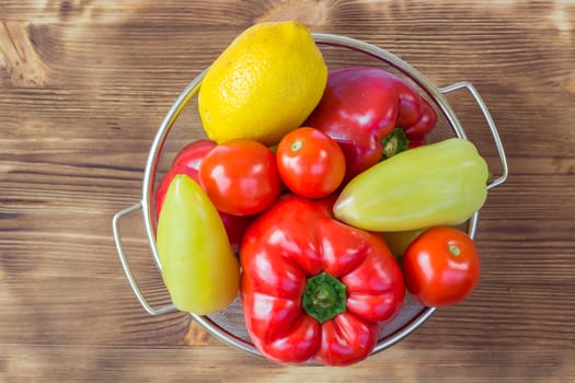 Vegetable still life of red and green peppers, tomatoes and lemon in sieve bowl on wooden board background