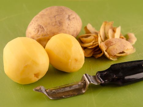 One unpeeled and two peeled potatoes on used green plastic board with peeler, simple food preparation illustration, vegetarian dieting, still life