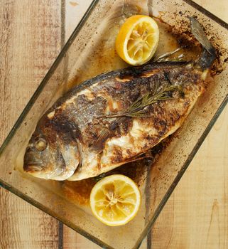 Delicious Homemade Roasted Sea Bream Fish with Lemons and Rosemary in Glass Cooking Tray closeup on Wooden background. Top View