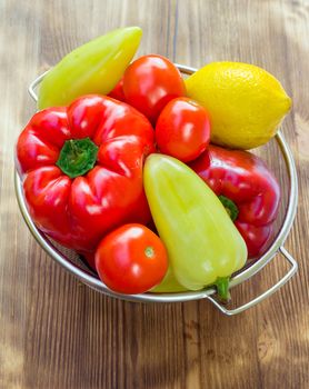 Vegetable still life of red and green peppers, tomatoes and lemon in sieve bowl on wooden board background