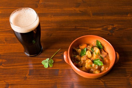 Traditional Irish Stew on a brown bowl with a pint of stout beer and a shamrock on a table.
