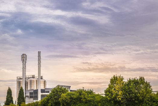 Image with industrial buildings, producer of  green electricity and heat, surrounded by nature, under a colorful sky.