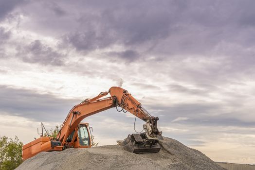 Heavy orange excavator with big bucket working on top of a ballast pile.