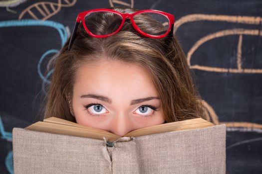 Perplexed  young student girl in red dress and red glasses reading a book hides behind it