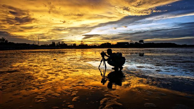 Silhouette of Asian photographer take photo with nature background, vibrant yellow of sky in sunset, wonderful landscape for Vietnam travel, tide going out at sea