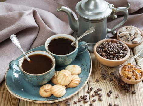 Two blue cups of black coffee, biscuits and coffee pot  surrounded by linen cloth, sugar pieces and coffee beans on old wooden table