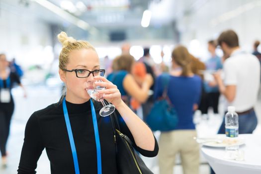 Business woman, wearing name tag, drinking glass of water during coffee break at business meeting or conference. Abstract blurred people socializing n background.