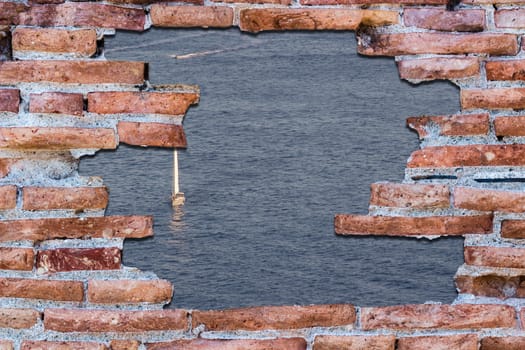 Ancient red brick wall with a hole over the blue sea with sailboat.