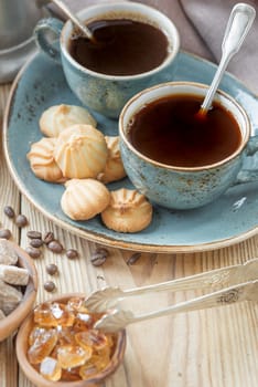 Two blue cups of black coffee, biscuits and sugar pieces surrounded by linen cloth and sugar-tongs on old wooden table