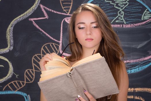 Young student girl in red dress and red glasses reads a book in front of school blackboard