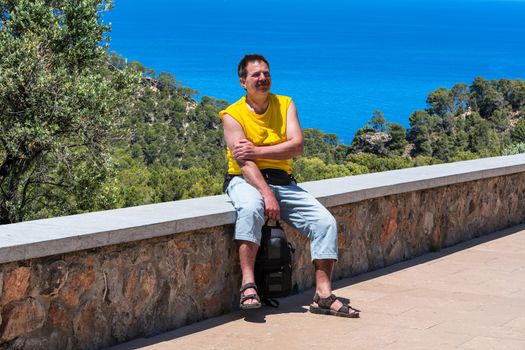 A man sitting in the sunshine on a wall in the background pine forest and the blue sea. Shot in Mallorca, Spain.