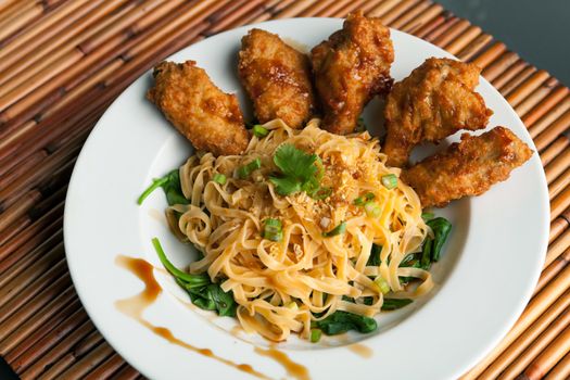 Thai style fried chicken wings on a round white plate with egg noodles and spinach.