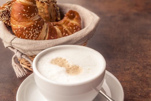 Delicious breakfast with a cup of milk coffee and fresh, sesame croissants in a rustic wicker basket, on a brown table.