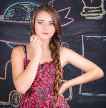 Young student girl in red dress poses in front of school classroom blackboard