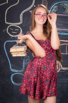 Young student girl in red dress with red glasses and pack of books poses in front of school classroom blackboard