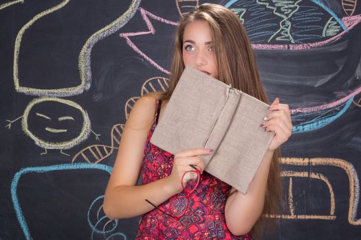 Perplexed  young student girl in red dress and red glasses reading a book hides behind it