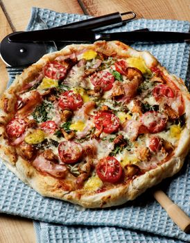 Delicious Mushrooms Pizza with Forest Chanterelles, Bacon, Tomatoes, Basil and Cheese on Blue Napkin with Pizza Cutter and Cutlery closeup on Wooden background