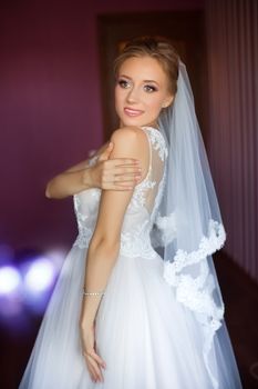 Portrait of beautiful bride with fashion veil posing on bed at wedding morning. Makeup. Brunette girl with long wavy hair styling. Wedding dress.